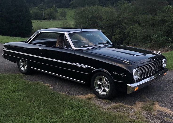 an old black car is parked on the side of the road in front of some trees