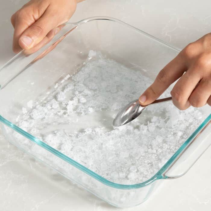 a person scooping sugar into a glass container with a spoon in it and ice cubes around the bowl