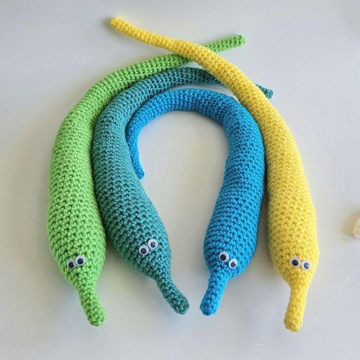 three crocheted bananas with googly eyes are on a white surface and one is green, the other is blue