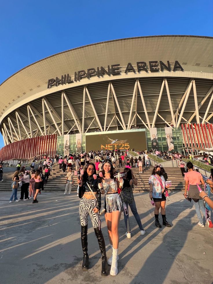 two women standing in front of a stadium with people walking around the building and onlookers