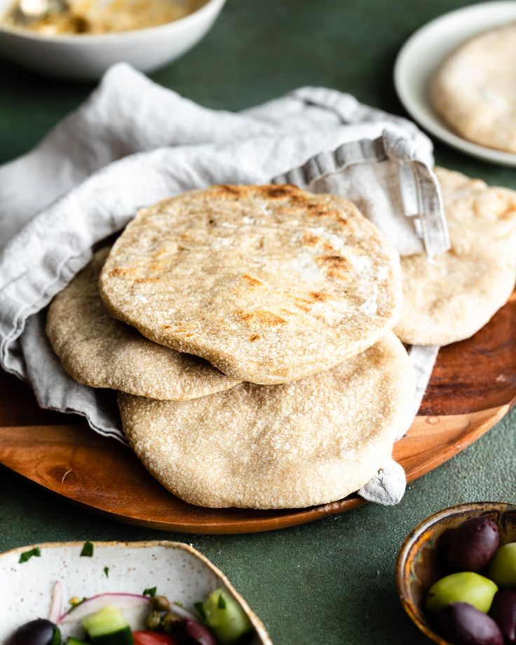 three pita breads sitting on top of a wooden plate with olives and cucumbers