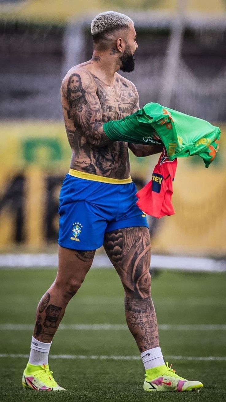 a tattooed man holding a green jacket on top of a soccer field with other players in the background