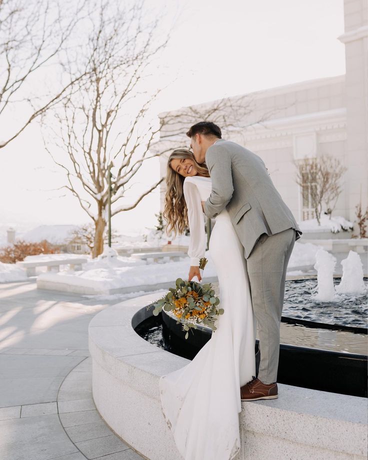 a bride and groom kissing in front of a fountain with flowers on it's side