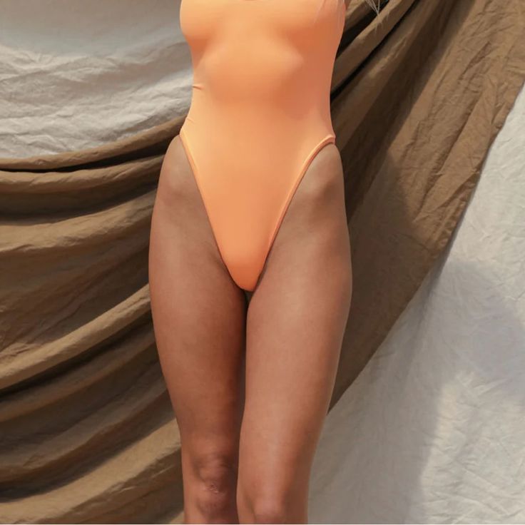 a woman in an orange one piece swimsuit standing next to a tan wall with her hands on her hips