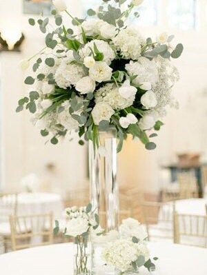 a tall vase filled with white flowers and greenery on top of a round table