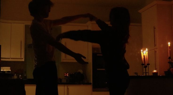 two people standing in a kitchen with candles on the counter and one person holding his arm out