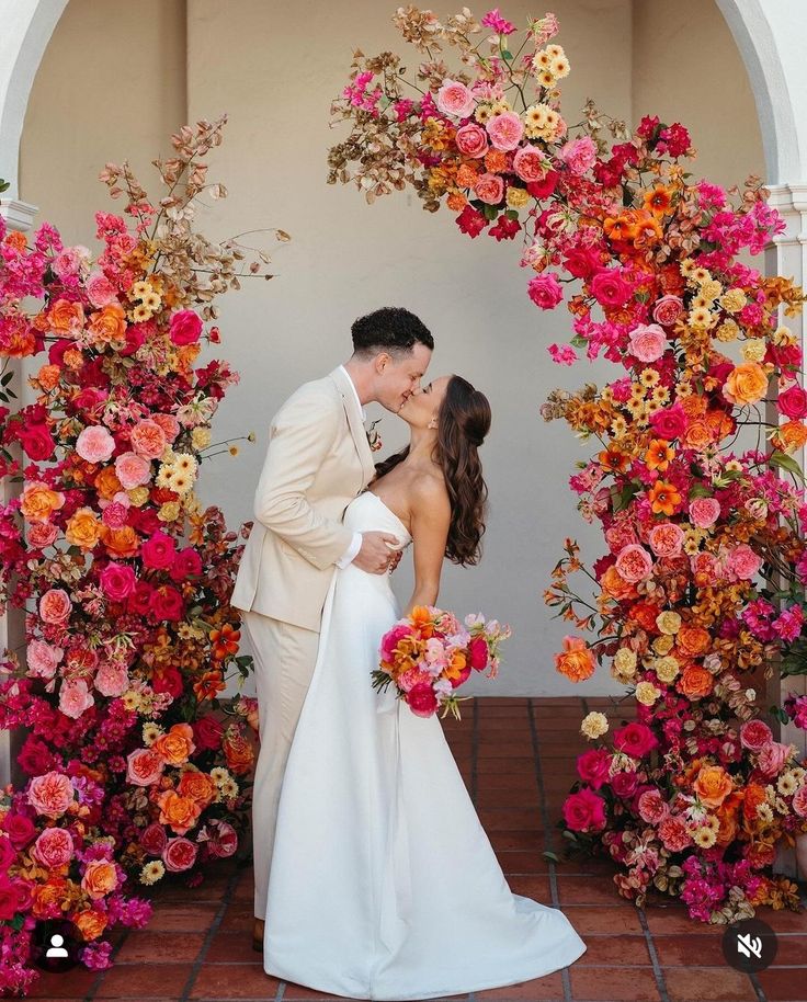 a bride and groom kissing in front of an archway with flowers on the wall behind them