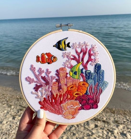 someone is holding up a colorful embroidery art piece on the beach with fish and corals