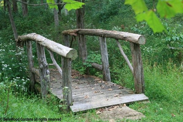 an old wooden bridge in the middle of some grass and trees with green leaves around it