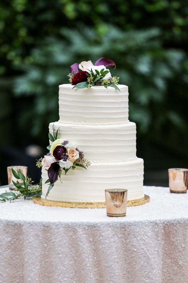 a three tiered white cake with flowers and greenery sits on top of a table