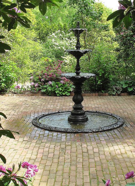 a water fountain in the middle of a brick walkway surrounded by trees and bushes with purple flowers around it