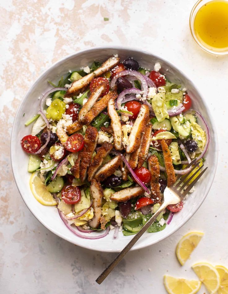 a salad with chicken, tomatoes, cucumber, onions and olives in a white bowl