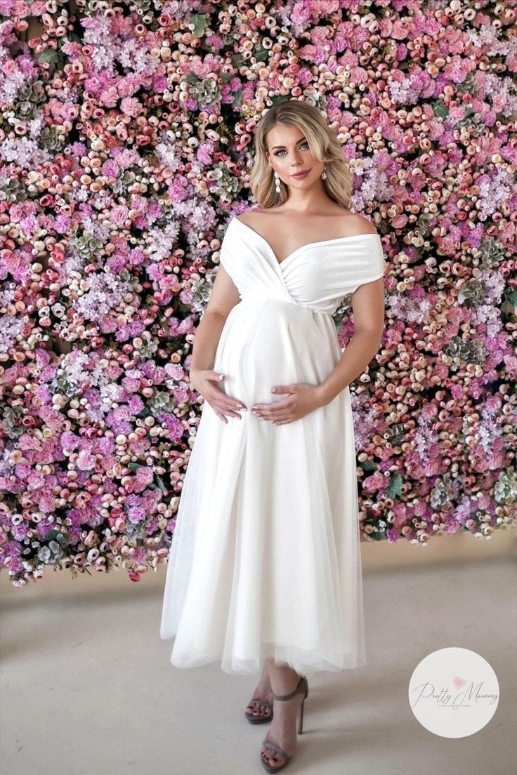 a pregnant woman wearing a white dress standing in front of a floral wall with pink flowers