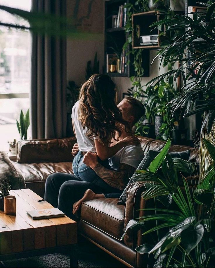 a man and woman sitting on a couch in front of a window with potted plants