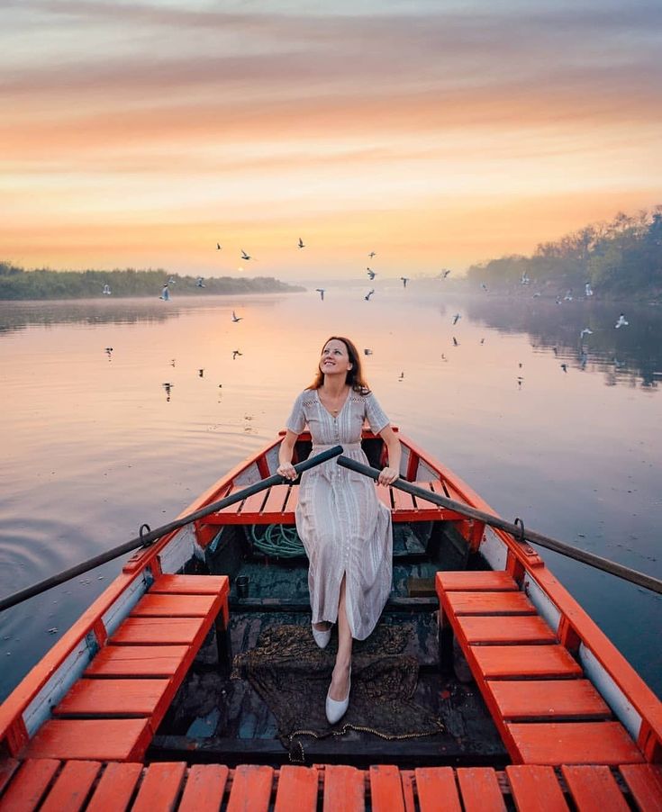 a woman sitting in the bow of a boat on a lake with birds flying around