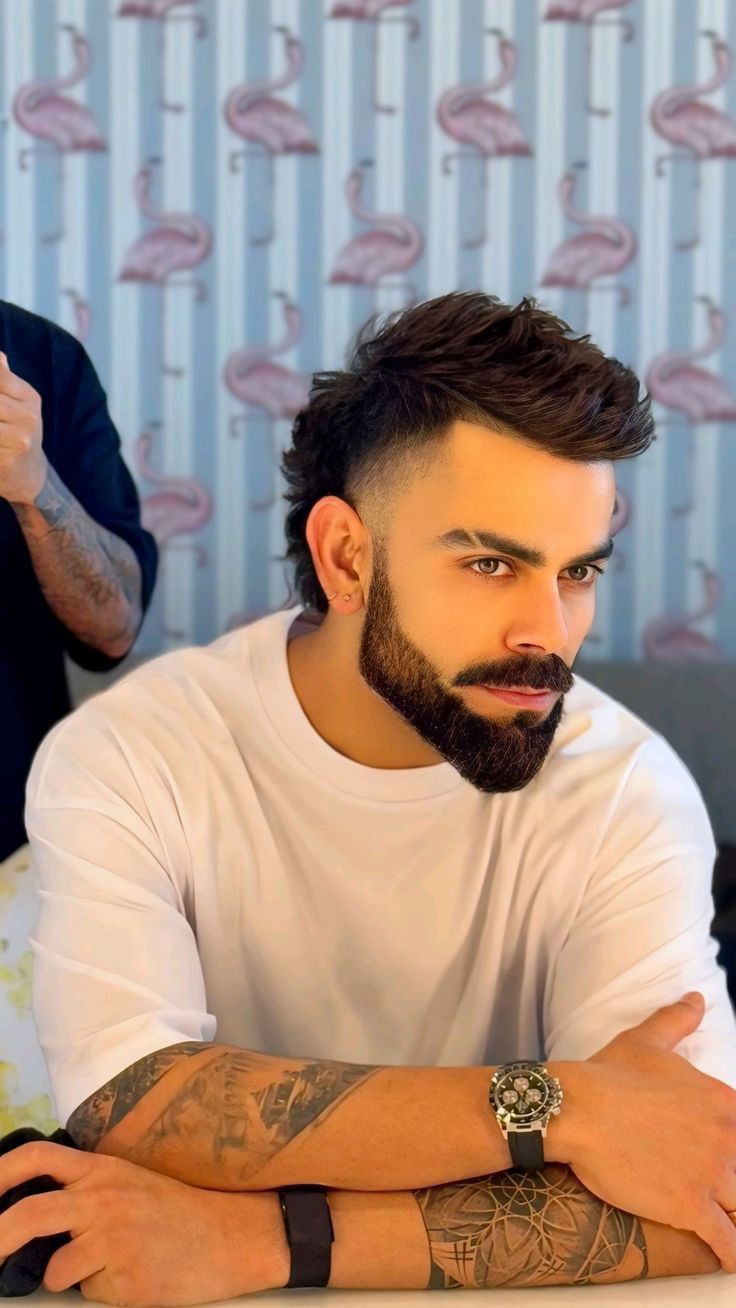 a man with a beard sitting at a table in front of a flamingo wallpaper