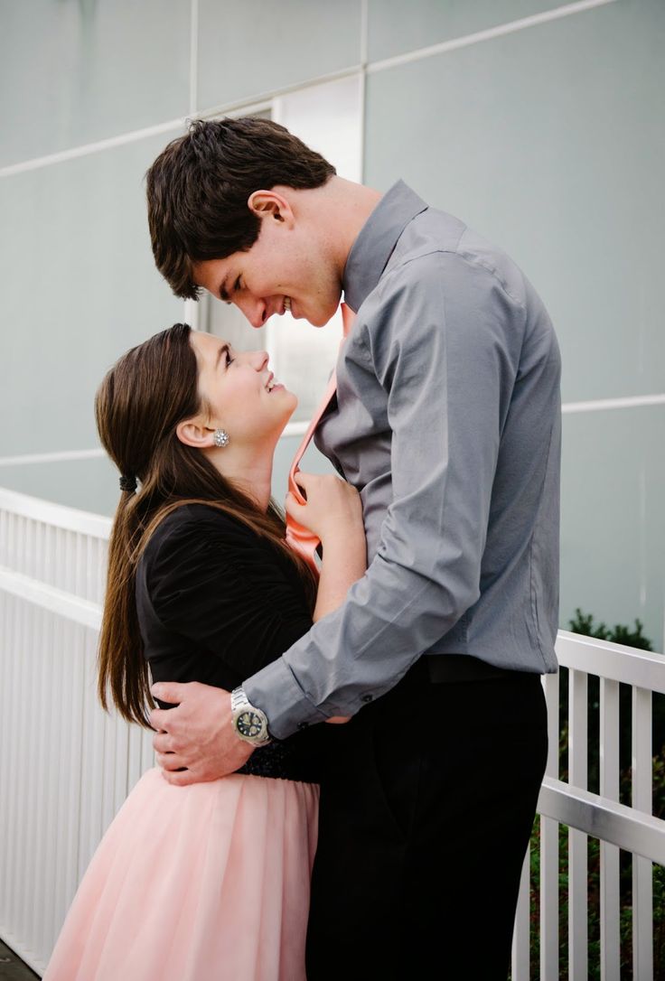 a man and woman standing next to each other in front of a white fence with their arms around each other