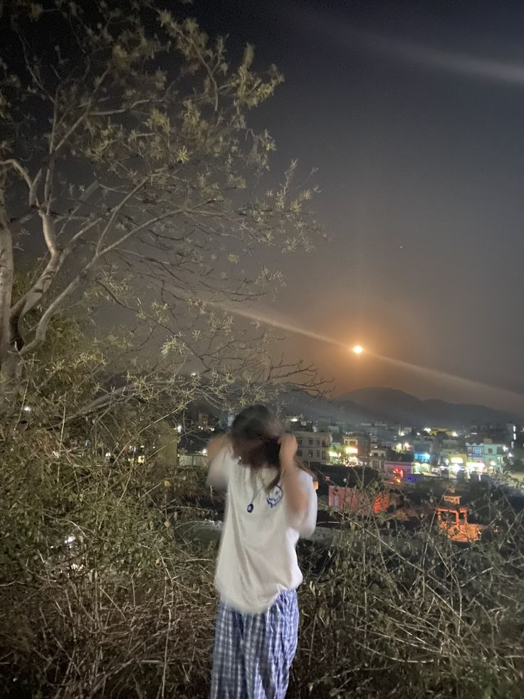 a person standing in front of a tree at night with the city lights behind them