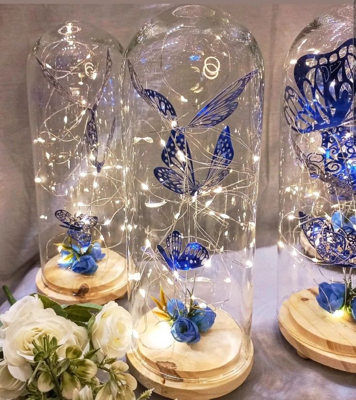 two glass domes with blue butterflies on them and white flowers in the bottom one is surrounded by fairy lights