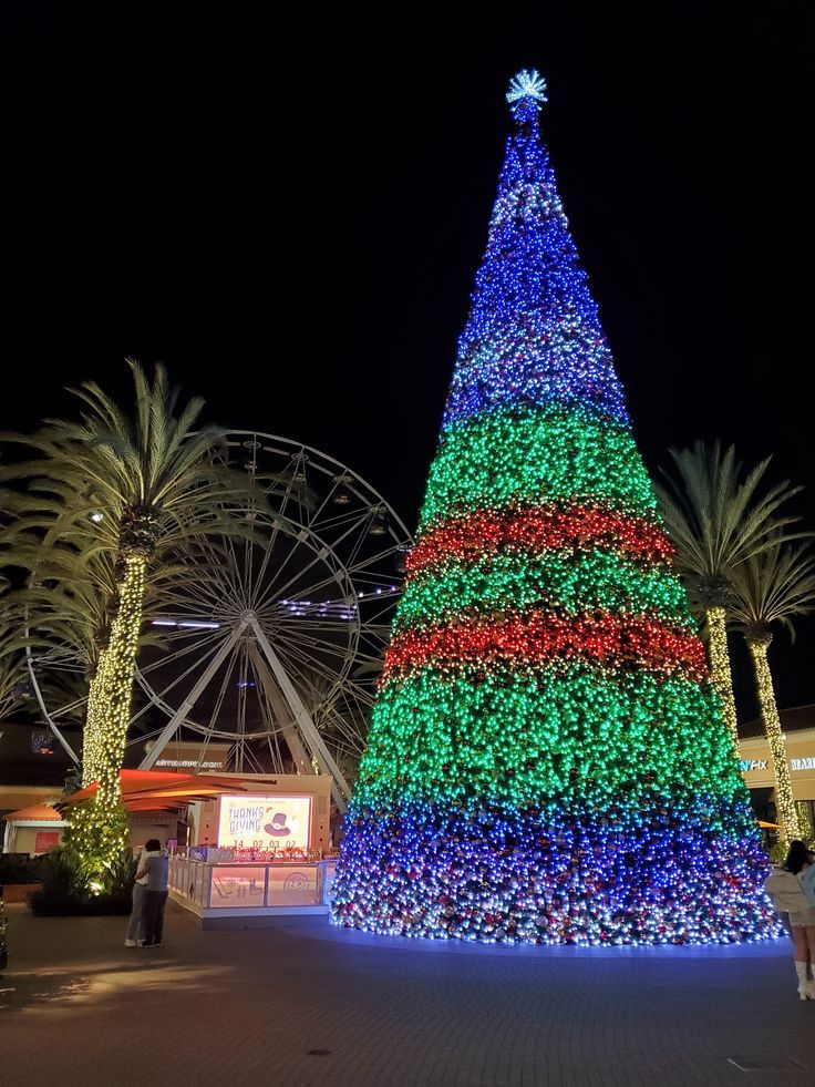 a brightly lit christmas tree in front of a ferris wheel and palm trees at night