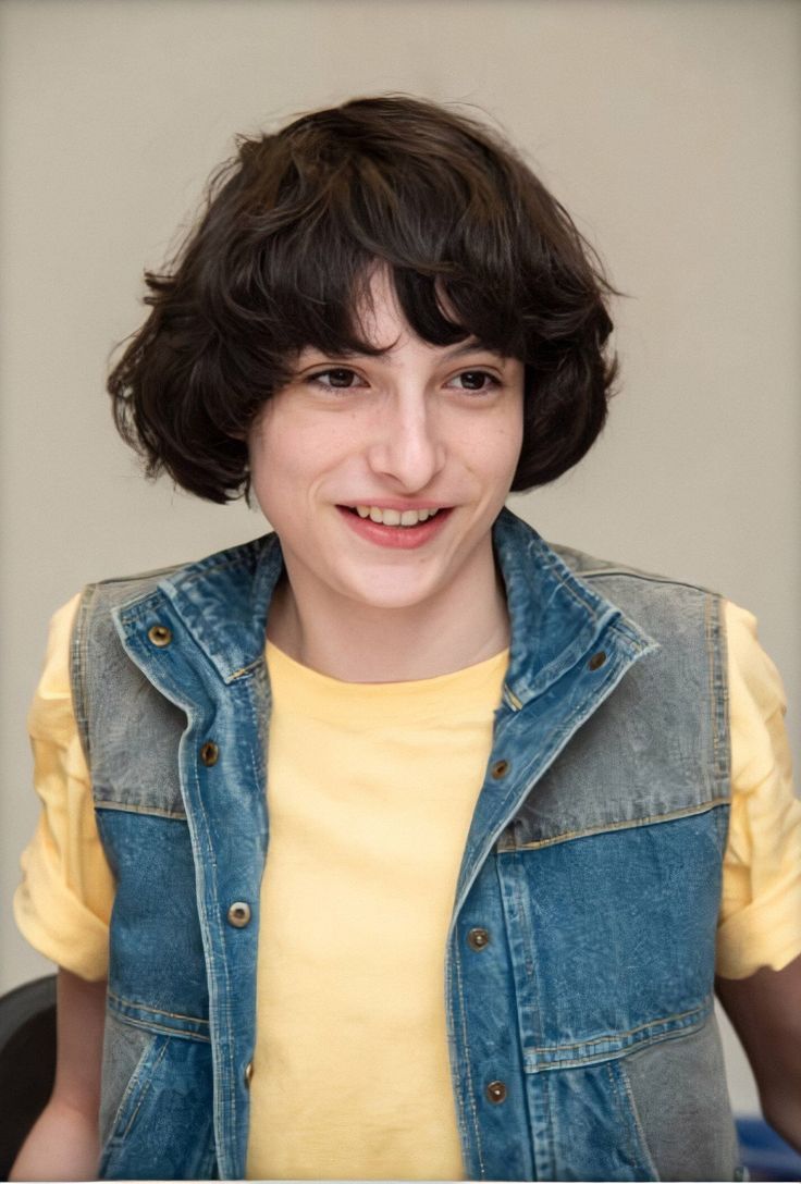 a young person with short hair and a denim vest smiling at the camera while wearing a yellow t - shirt