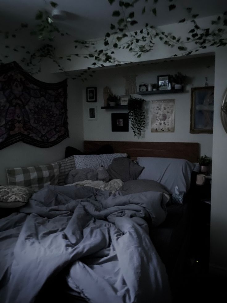 an unmade bed in a dark room with lots of plants hanging from the ceiling