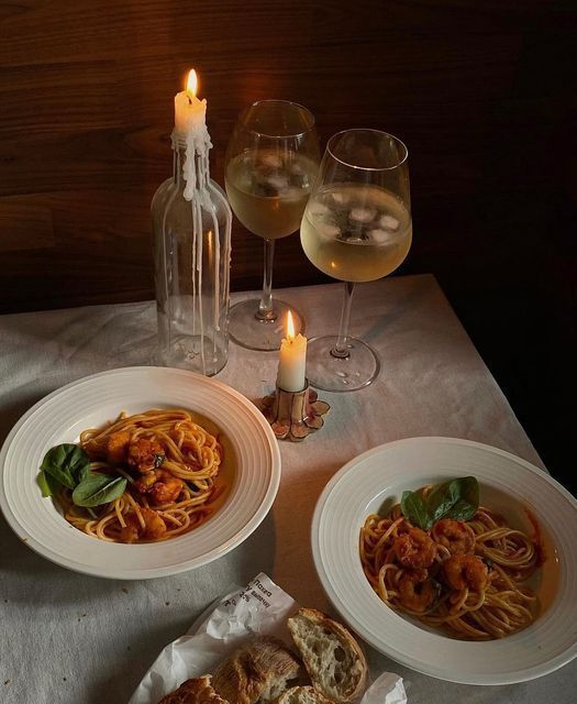 two plates of pasta and wine on a table with candles in the background at night