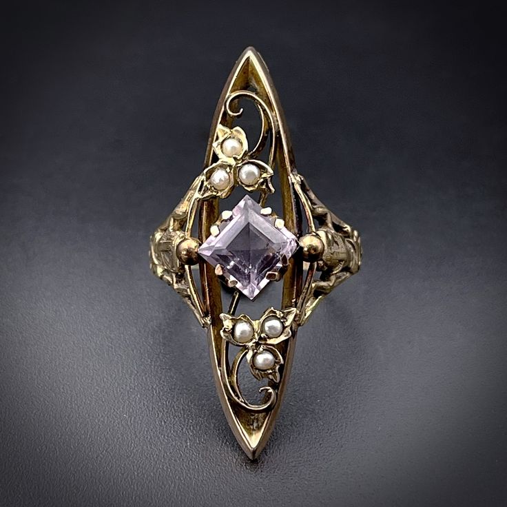 "Have you heard the saying, \"What's Old is New\"? Here we have just the example! Converted from an antique brooch into this stunning statement ring, this piece embraces the Art Nouveau sensibility with flowing lines and an homage to  Mother Nature. Golden three petaled flowers with curly vines are separated by a diamond shaped set amethyst stone as the central focus. Seed pearls are set in the petals of the flowers.  The swirling 9 karat gold framework  works itself in and around the two blossoms, creating a sense of movement and interest on the ring top.  But that's not all!  The shank is a stunning Egyptian revival replica of a previously owned Art Deco ring depicting the Egyptian Goddess, Nut, in a crouched position, holding up the sky. She appears on both shoulders of the shank and is Antique Amethyst Ring For Formal Occasions, Vintage Amethyst Ring With Intricate Design, Victorian Amethyst Ring For Anniversary, Antique Gold Amethyst Ring, Victorian Style 14k Stamped Amethyst Ring Gift, Victorian Style Amethyst Ring Stamped 14k, Vintage Amethyst Ring With Rose Cut Diamonds, Antique Amethyst Ring With Intricate Design, Antique Amethyst Ring With Rose Cut Diamonds For Wedding