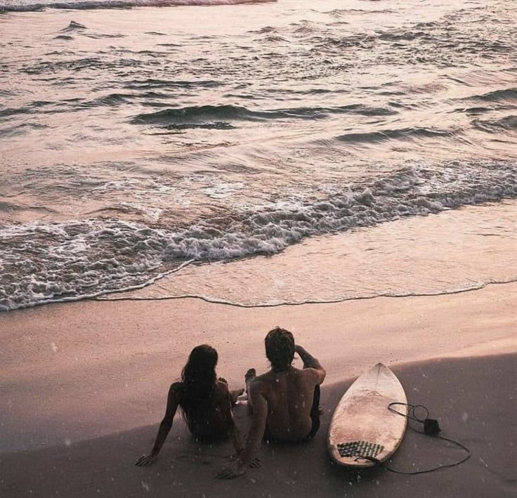 two people sitting on the beach next to a surfboard looking out at the ocean