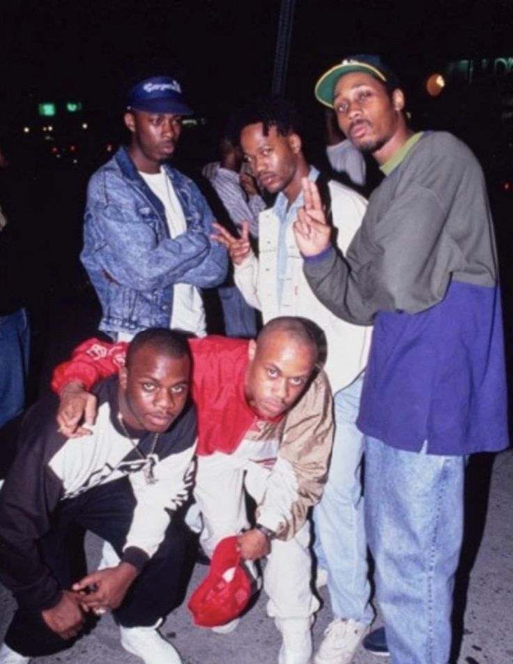 four men posing for the camera in front of a building at night with their arms around each other