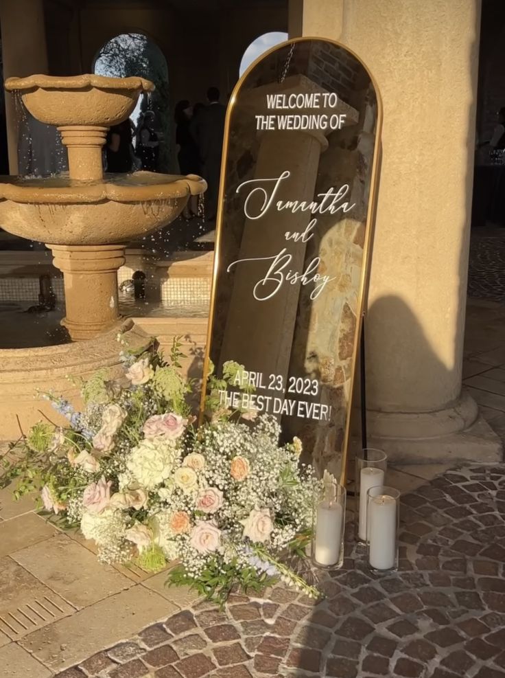 a welcome sign with flowers and candles in front of a fountain on a brick walkway