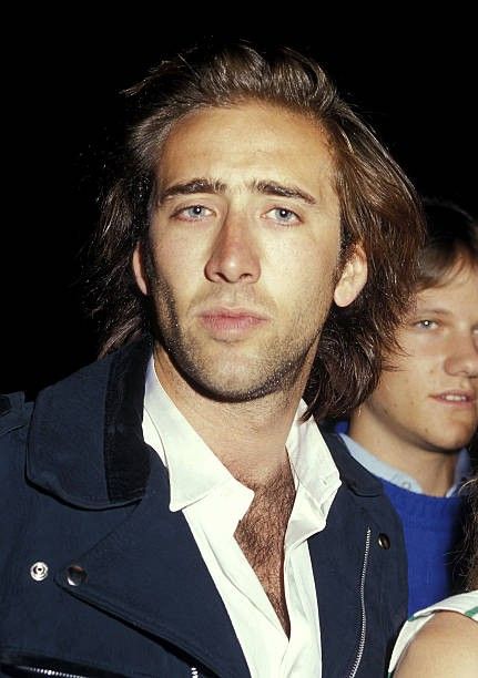 a man with long hair standing next to a woman in a white shirt and black jacket