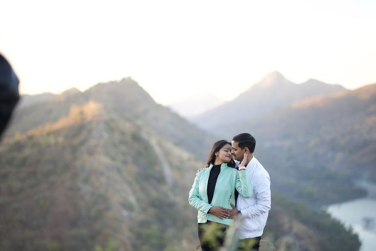 a man and woman standing next to each other on top of a hill with mountains in the background