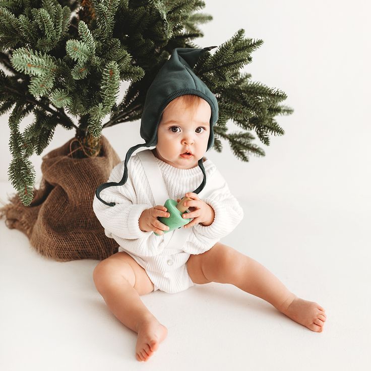 a baby is sitting next to a christmas tree and holding a cup in his hands