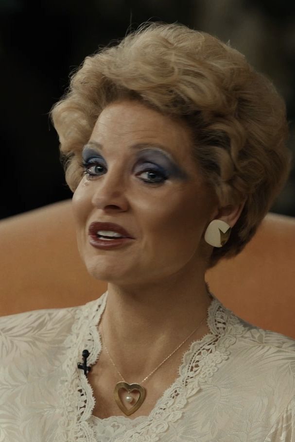 an older woman with blue eyes and makeup looks at the camera while sitting in a chair