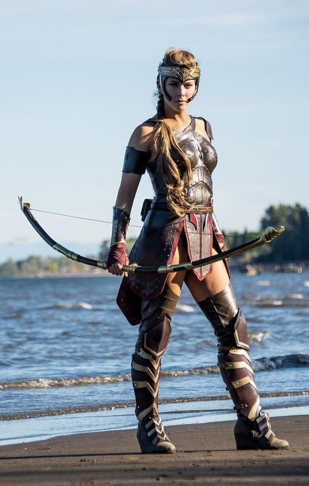 a woman dressed in armor holding a bow and arrow while standing on the beach next to the water