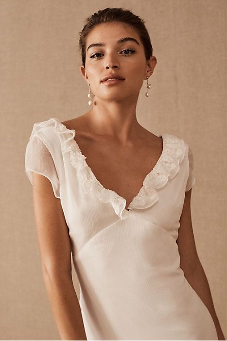 a woman wearing a white top with ruffles on the neck and shoulders, standing in front of a beige background