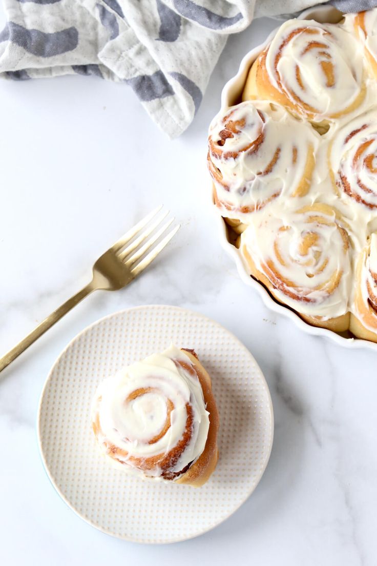 a plate with cinnamon rolls on it next to a bowl of icing and a fork