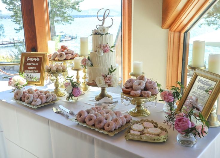 a table topped with lots of donuts covered in frosting and pink flowers next to a window