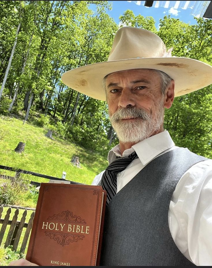 an older man wearing a cowboy hat and holding a bible in his hand while standing outside