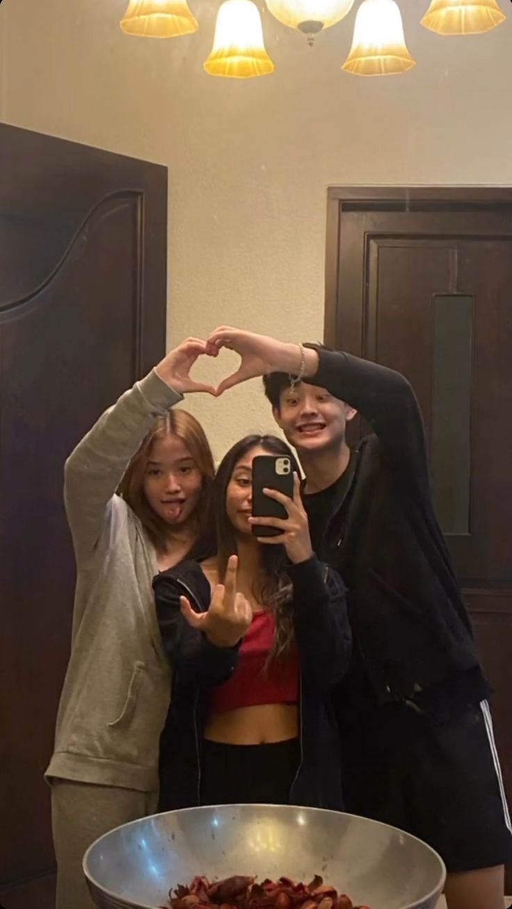 three people standing in front of a cake making a heart shape with their hands while taking a selfie