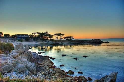 the sun is setting over the water and rocks on the shore with trees in the background