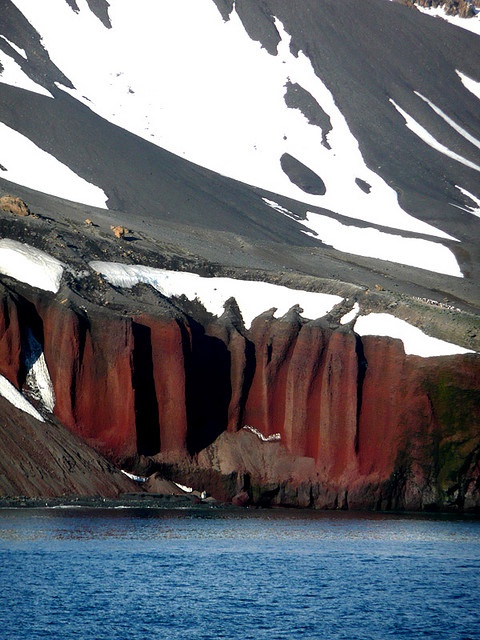 there is snow on the mountains and water in the foreground, with red cliffs to the right