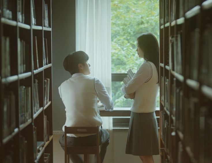 two people sitting at a table in front of bookshelves and looking out the window