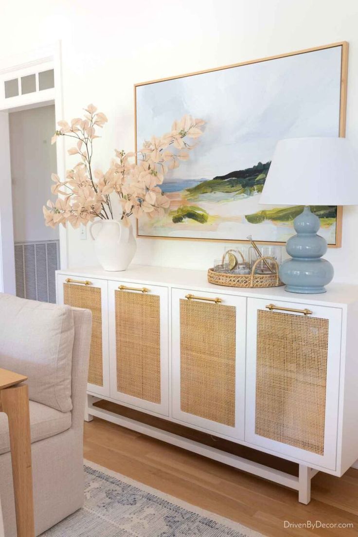 a white sideboard with wicker doors and flowers in a vase on top, next to a beige chair