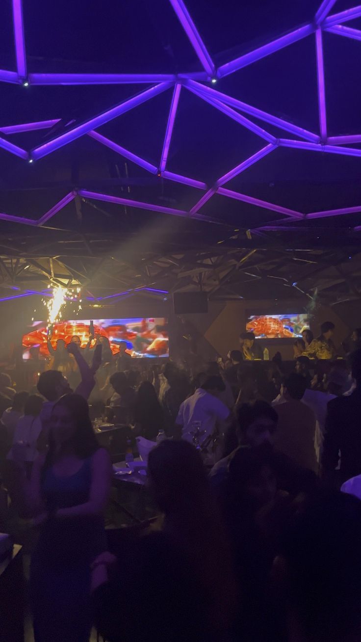 a group of people standing around in a room with purple lights on the ceiling and televisions above them