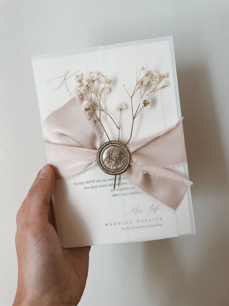 a hand holding a wedding card with flowers on the front, and a ribbon tied around it