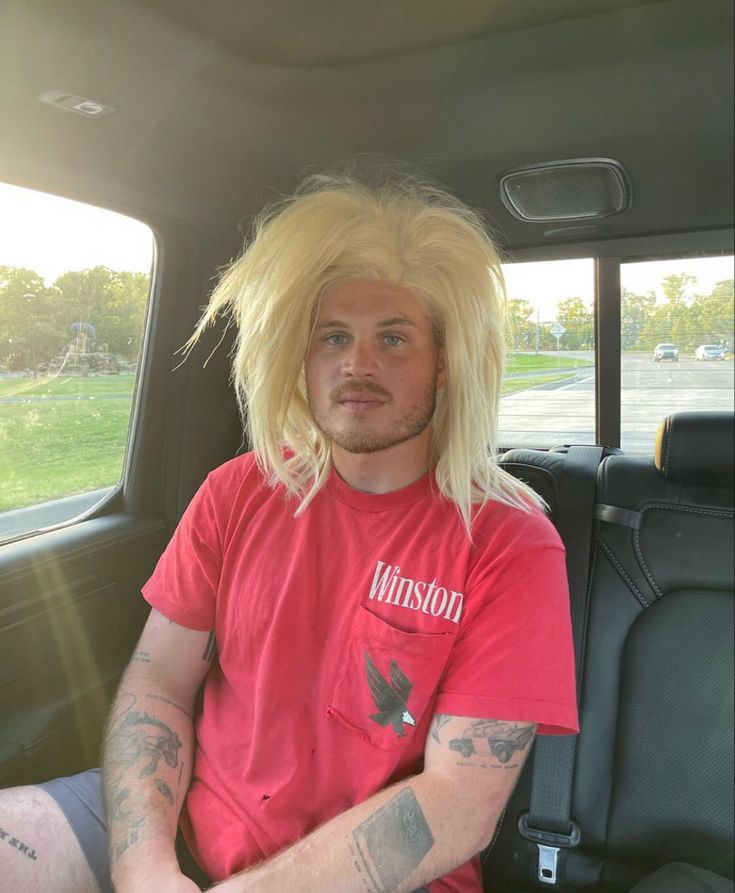 a man with blonde hair and tattoos sitting in the back seat of a car wearing a red shirt