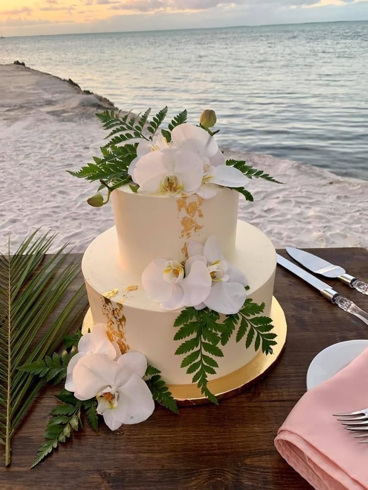 a wedding cake with white flowers and greenery sits on a table next to the ocean