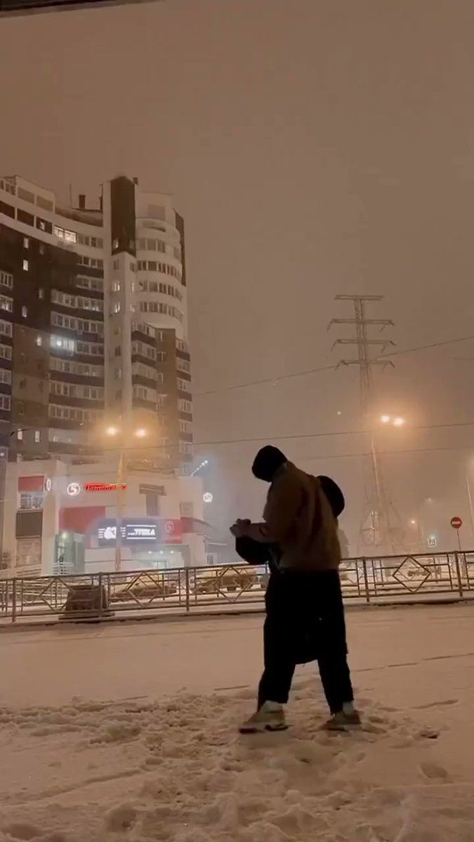 a man walking across a snow covered street in front of some tall buildings at night